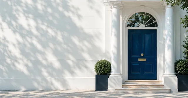 A navy Georgian front door with a fanlight and white stucco portico, with dappled tree shadows on a wide white wall to the left