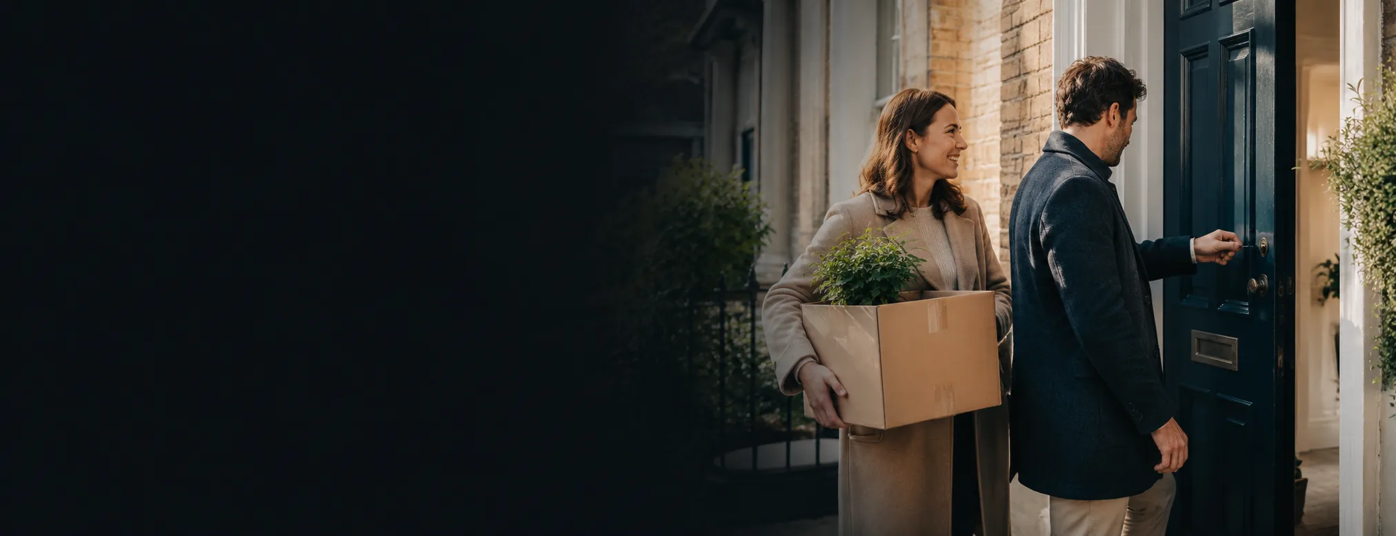 A couple carrying a box and a plant unlocking the navy front door of their new home